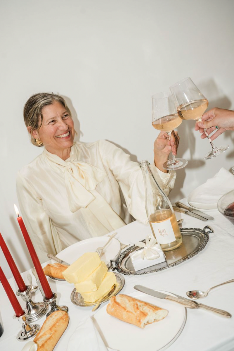 Melinda at a dining table with Lorenza Rosé Bubbly, wine glasses and bread, smiling.