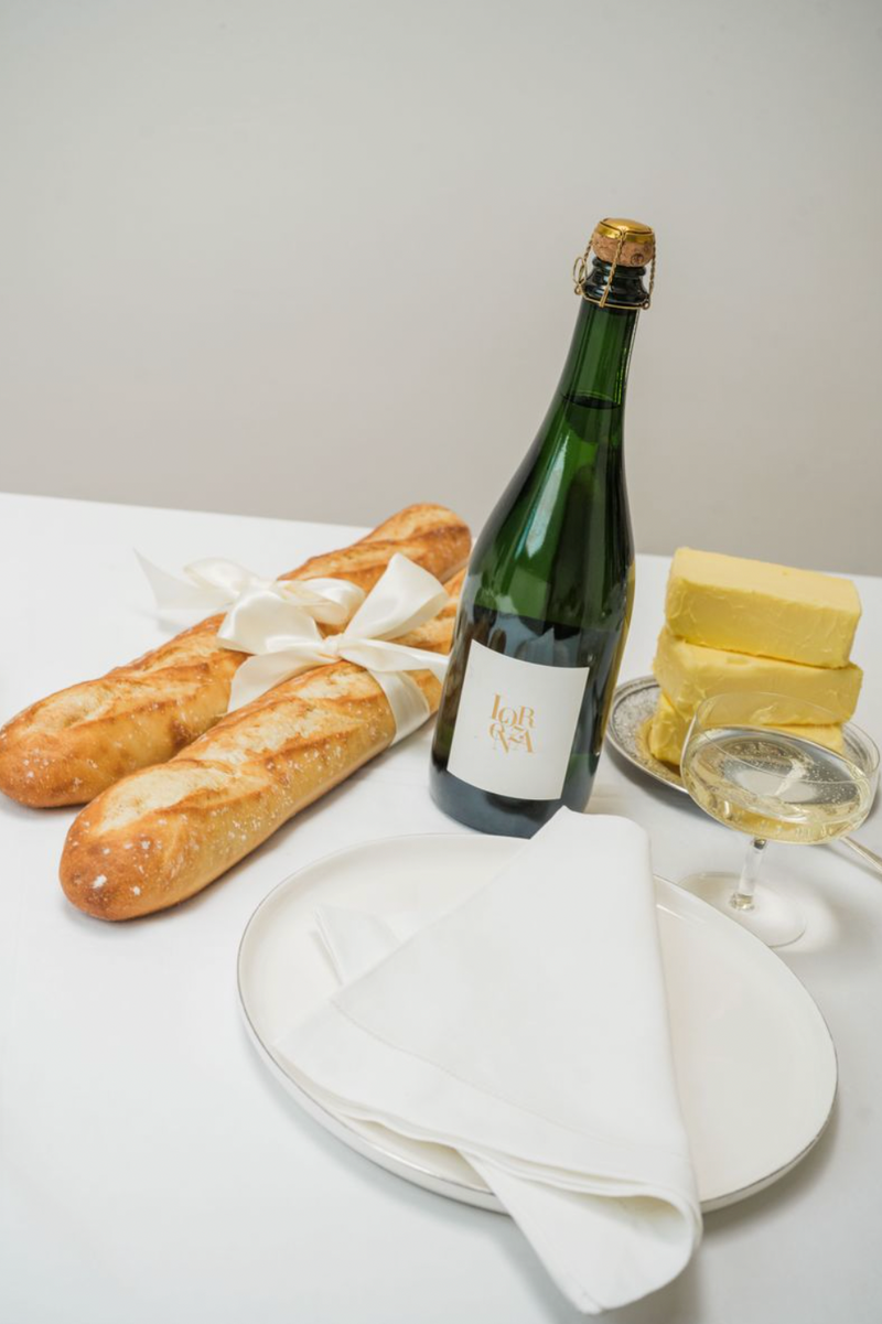 Bottle of Lorenza Sparklng Picpoul Blanc, two baguettes, and cheese on a white tablecloth.